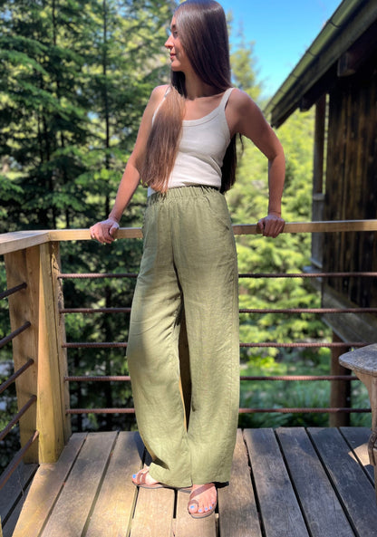 A woman stands on a wooden deck outdoors, wearing green linen pants, a white tank top, and brown sandals, with trees and a cabin in the background.