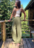 A woman stands on a wooden deck outdoors, wearing green linen pants, a white tank top, and brown sandals, with trees and a cabin in the background.