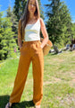 A woman standing outdoors on a grassy lawn wearing cedar linen pants, a white tank top, and sandals, with trees and blue sky in the background.
