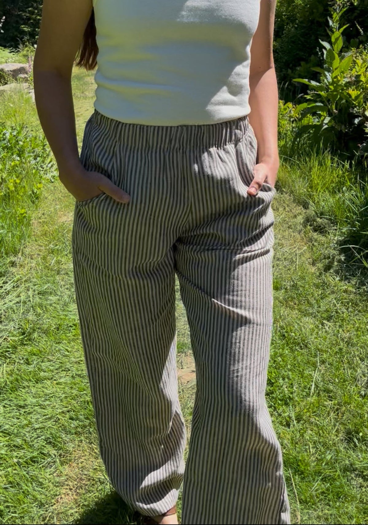 A woman wearing a white tank top and gray-and-white vertically striped linen pants, standing outdoors on a grassy path with green foliage in the background.