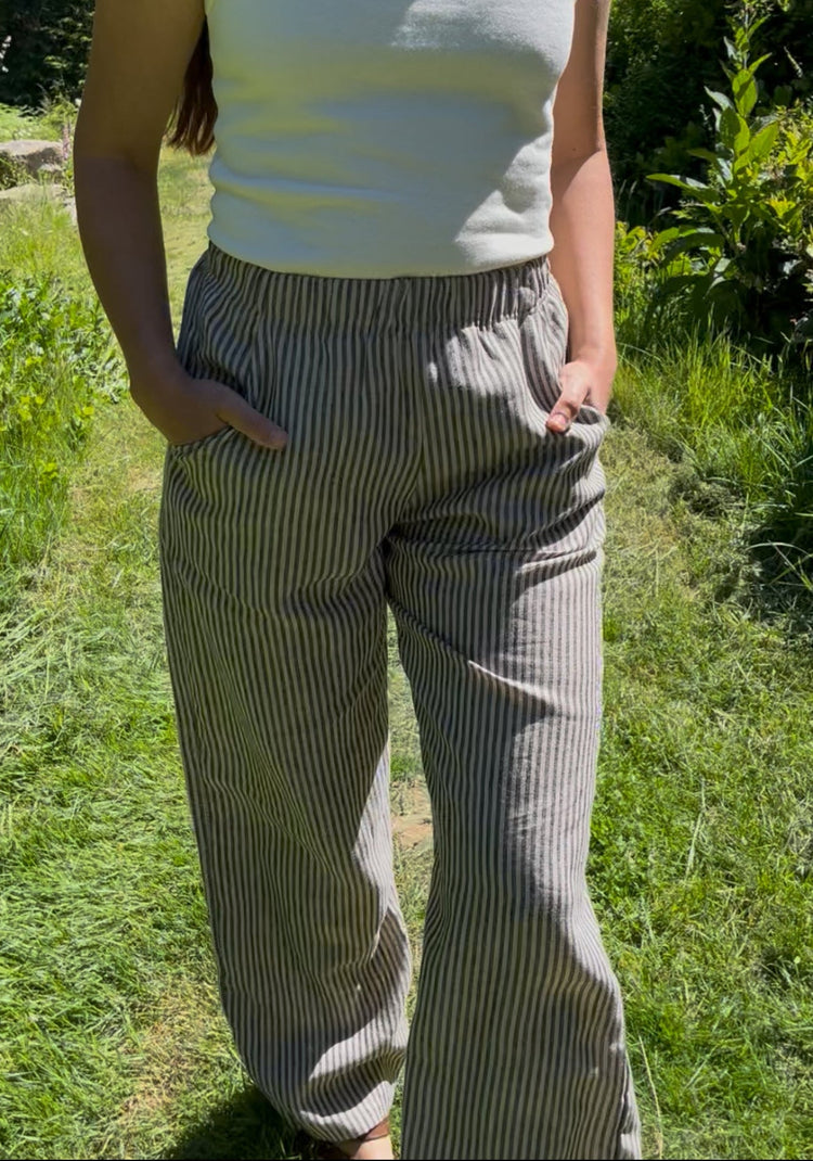 A woman wearing a white tank top and gray-and-white vertically striped linen pants, standing outdoors on a grassy path with green foliage in the background.