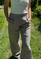 A woman wearing a white tank top and gray-and-white vertically striped linen pants, standing outdoors on a grassy path with green foliage in the background.