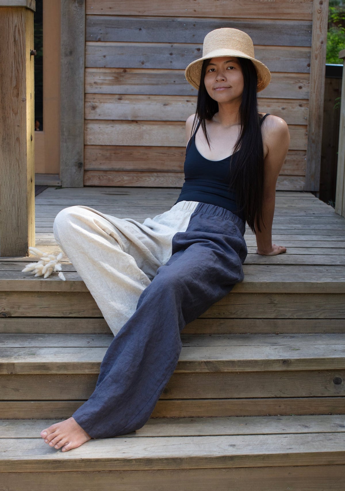 A woman sitting on wooden steps wearing two-toned linen lounge pants. One leg is dark grey, the other is light beige.