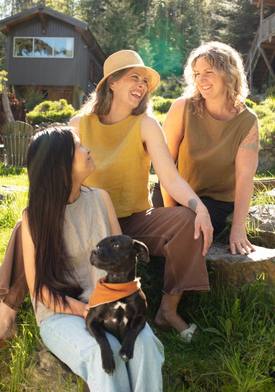 Three women sitting on a log wearing linen tops with a dog in a natural setting